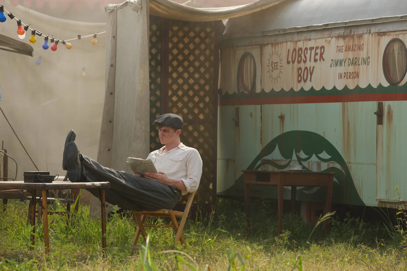 Evan Peters as Jimmy Darling reads beside a trailer painted with the words Lobster Boy and Jimmy Darling.