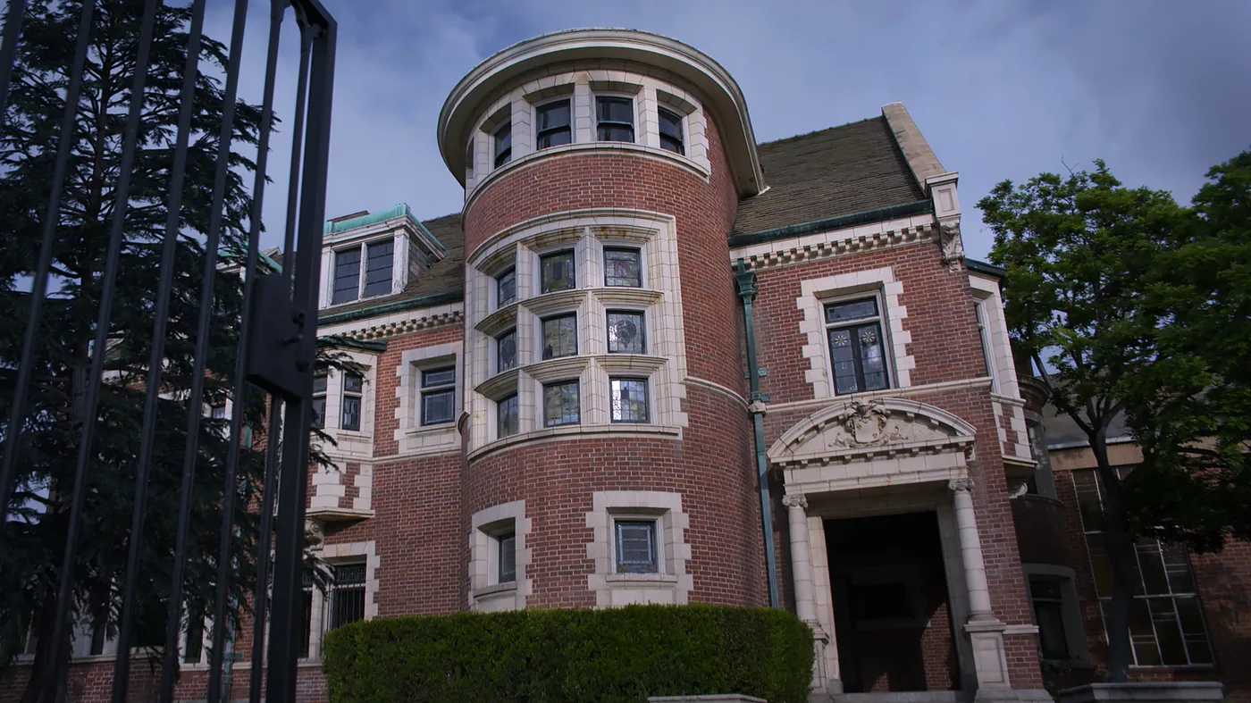 A brick house with a rounded tower appears behind a gate in an official American Horror Stories publicity still.