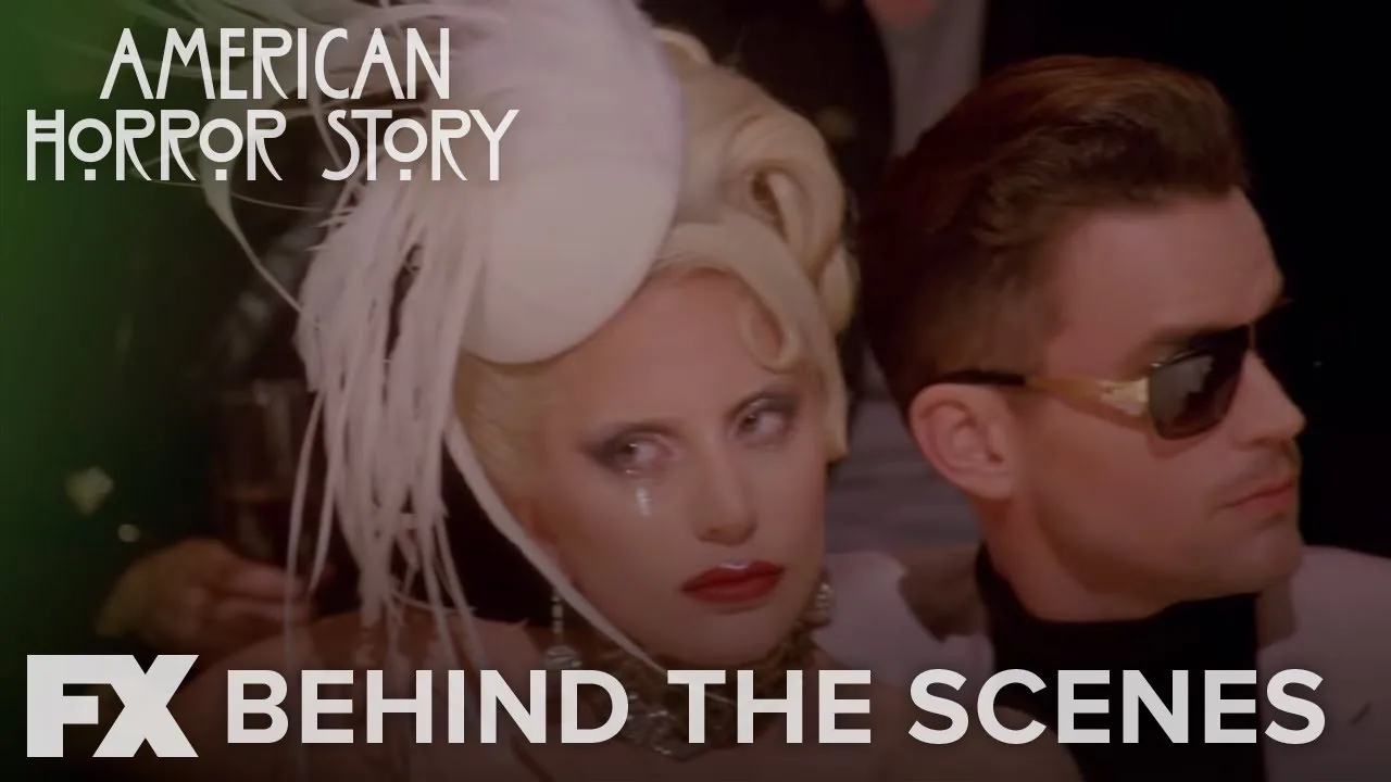 Lady Gaga as the Countess wears a white feathered headpiece beside a man in sunglasses in FX's official Hotel wardrobe video thumbnail.