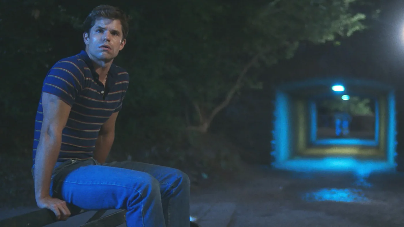 Charlie Carver as Adam sits outdoors at night in a striped polo shirt beside a blue-lit path.