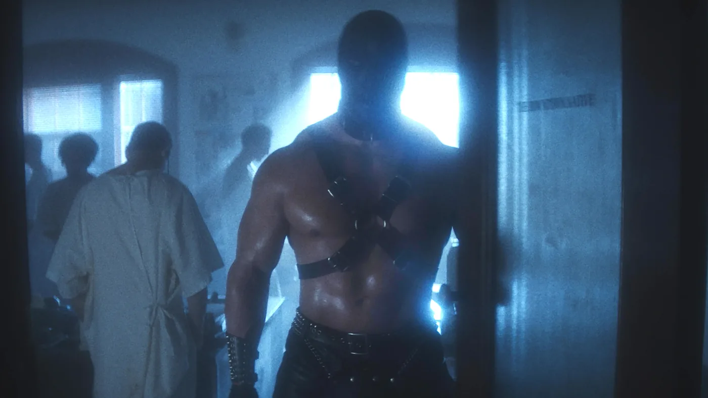 A broad-shouldered masked figure in a black chest harness stands in a blue-lit hospital corridor in a NYC publicity still.