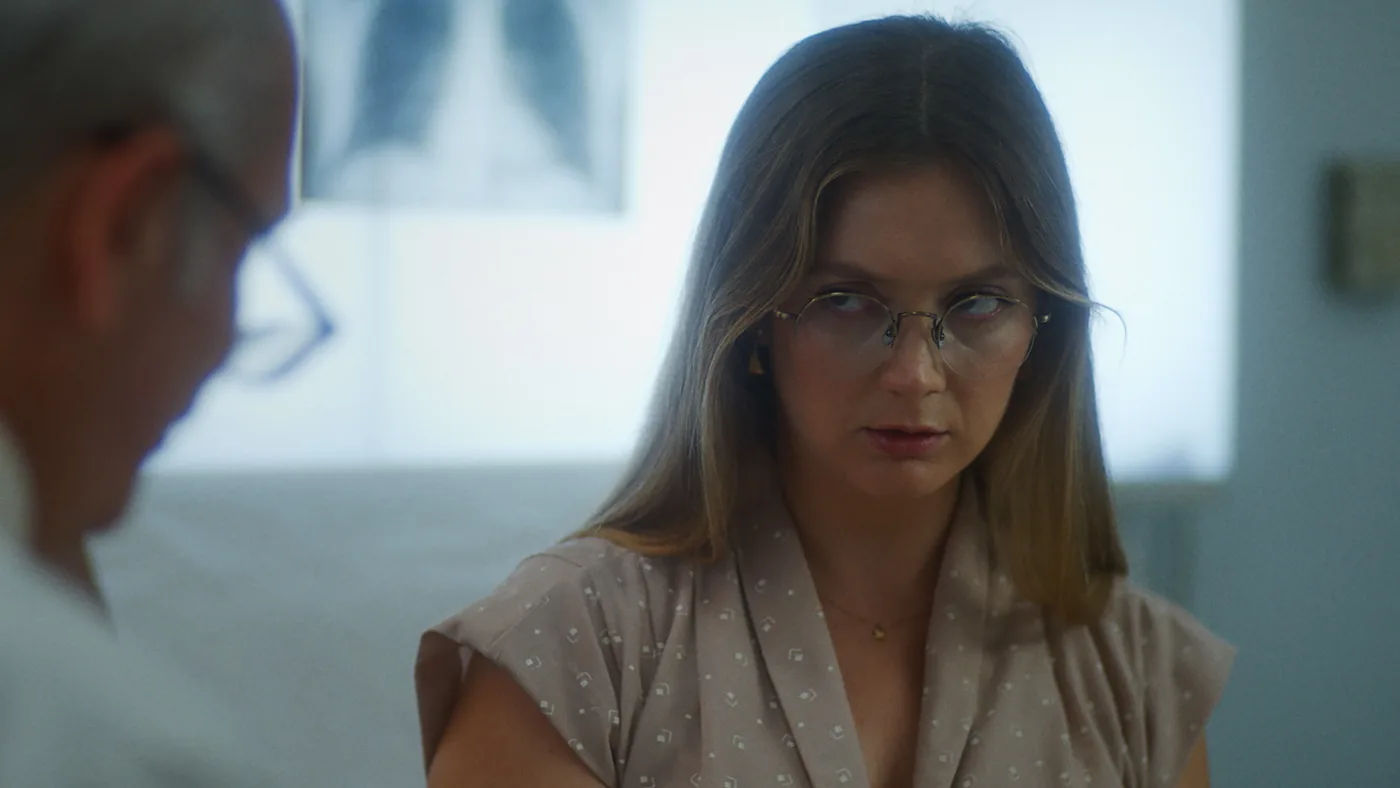 Billie Lourd as Hannah, wearing glasses and a pale blouse, looks toward another person in a doctor's office.