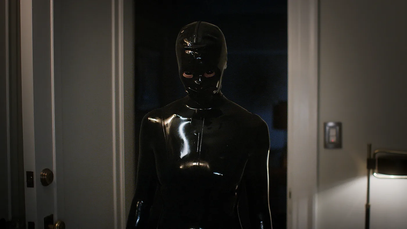 A figure in a black, glossy full-body rubber suit stands in a doorway in an official American Horror Stories publicity still.