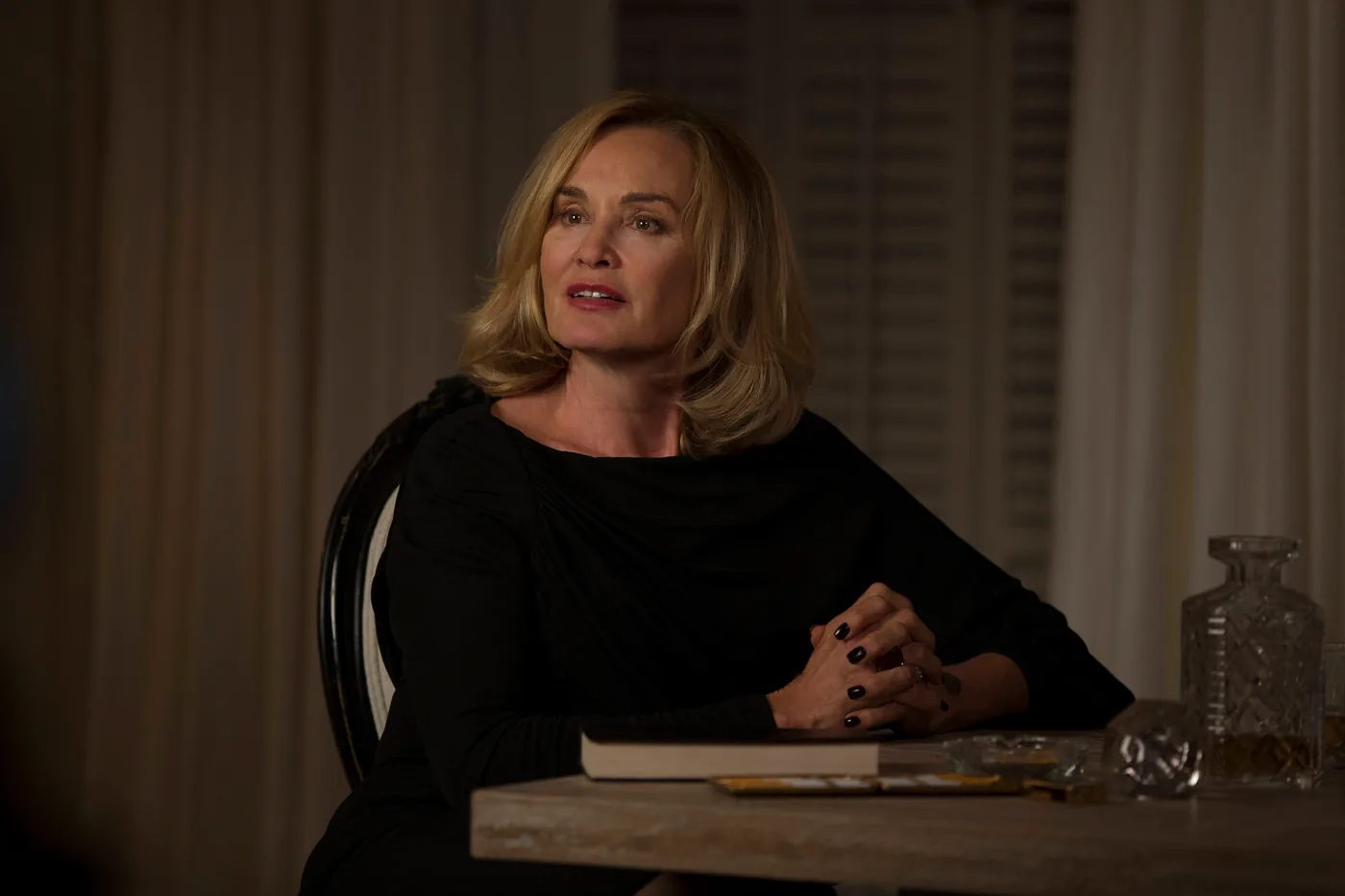 Jessica Lange as Fiona Goode sits indoors at Miss Robichaux's Academy wearing black in a Coven publicity image.