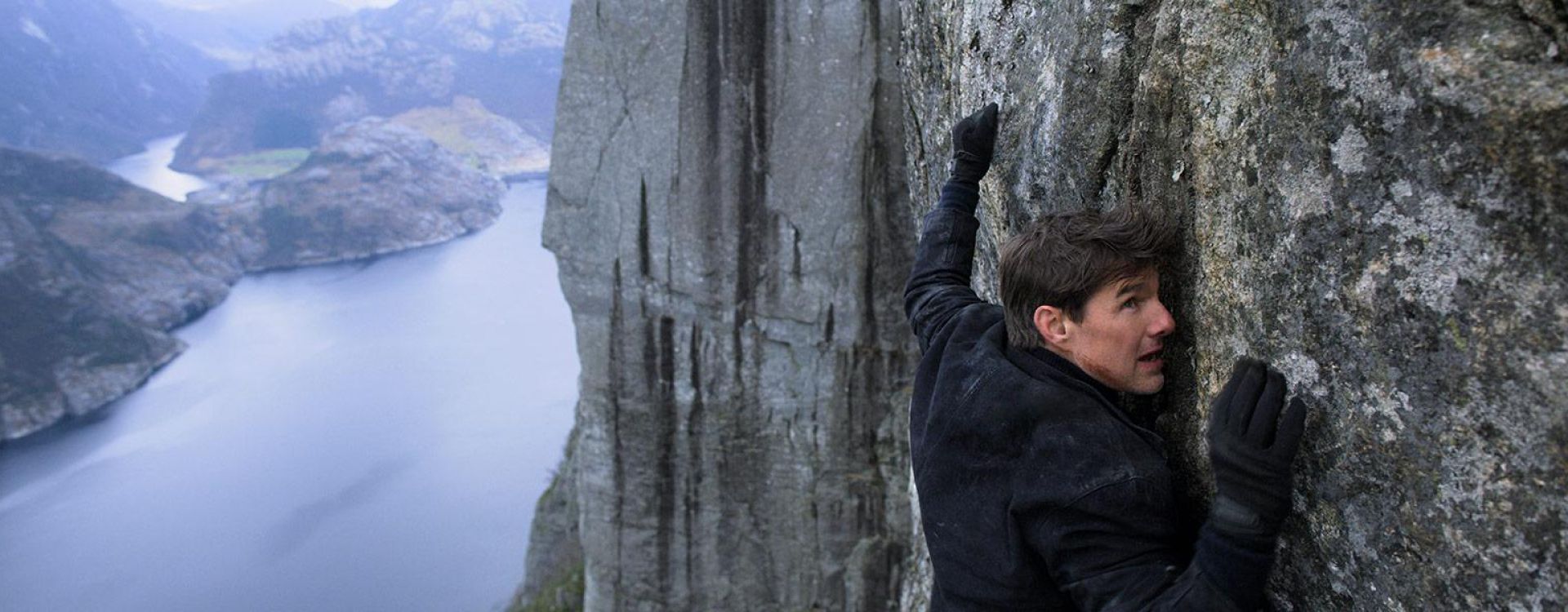 Tom Cruise as Ethan Hunt clinging to a cliff in official Mission: Impossible – Fallout art.