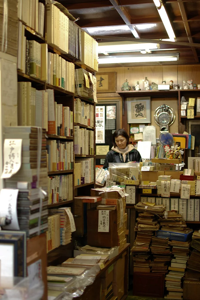 Bookshelves and stacks of old books inside a used bookstore in Jinbocho, Tokyo, with a person behind the counter.