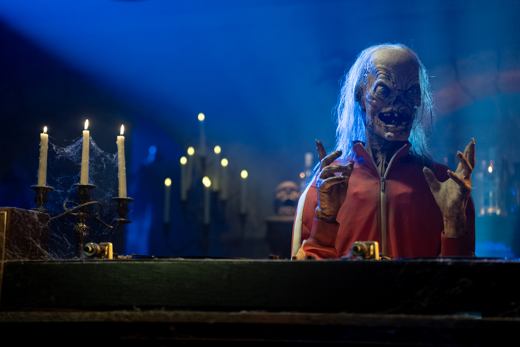 The rebuilt Crypt Keeper puppet in a red outfit behind a desk, lit in blue, in AMC's 2026 FearFest first-look photo.