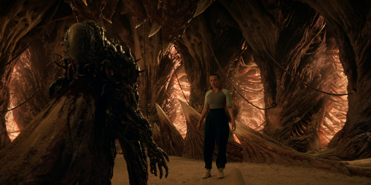 Millie Bobby Brown as Eleven standing before a towering fleshy creature inside the Upside Down in Stranger Things 5