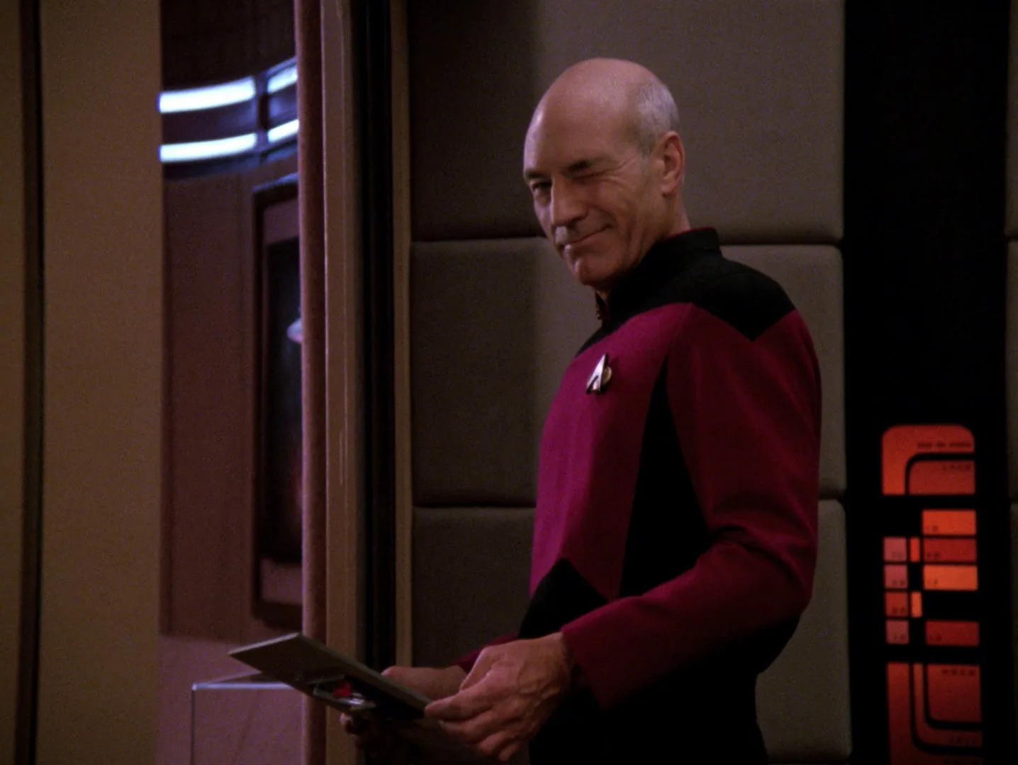 Captain Picard smiling slightly while holding a data pad in a corridor aboard the Enterprise in the TNG episode Disaster.