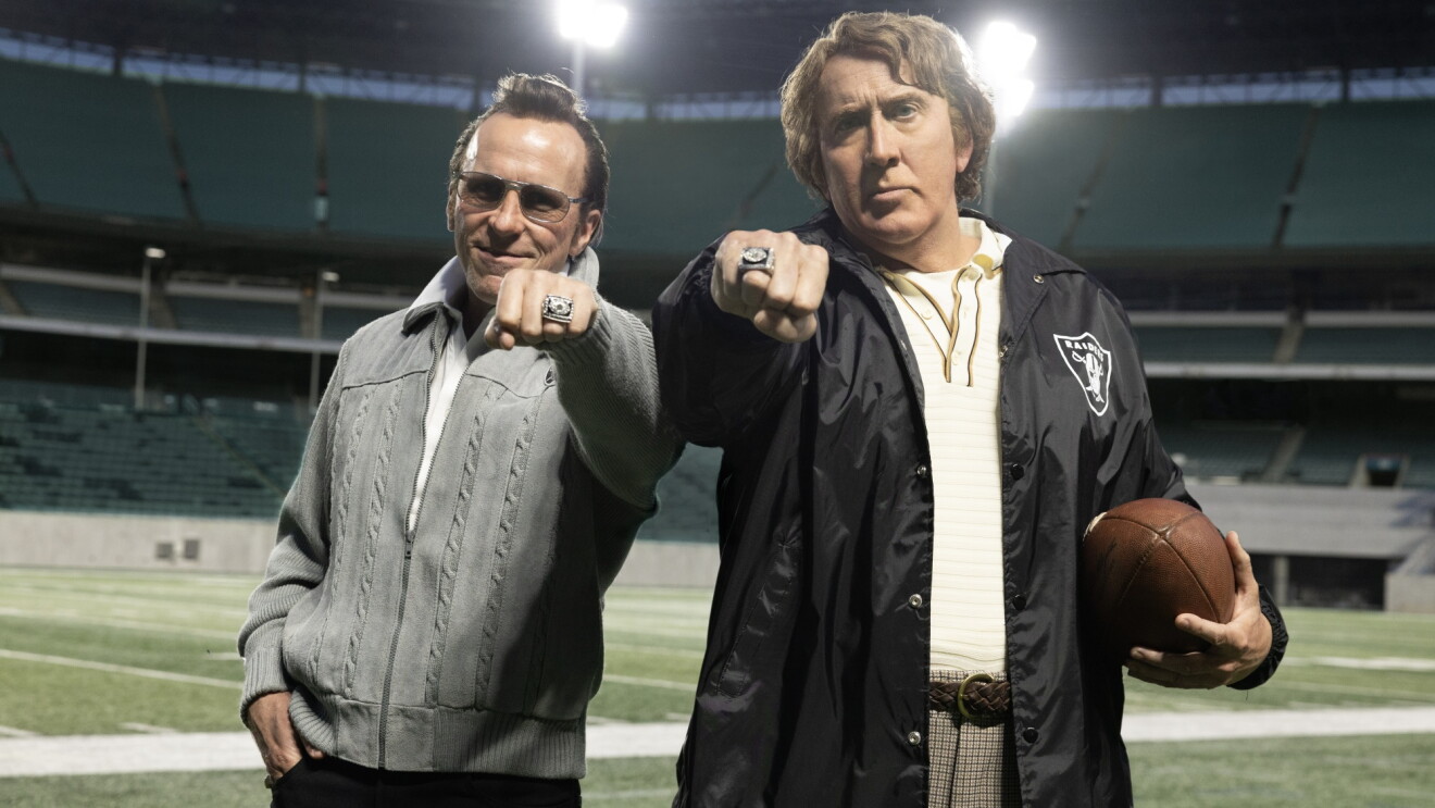 Christian Bale as Al Davis and Nicolas Cage as John Madden standing on an empty football field wearing Super Bowl rings, with Cage holding a football and wearing a Raiders jacket.