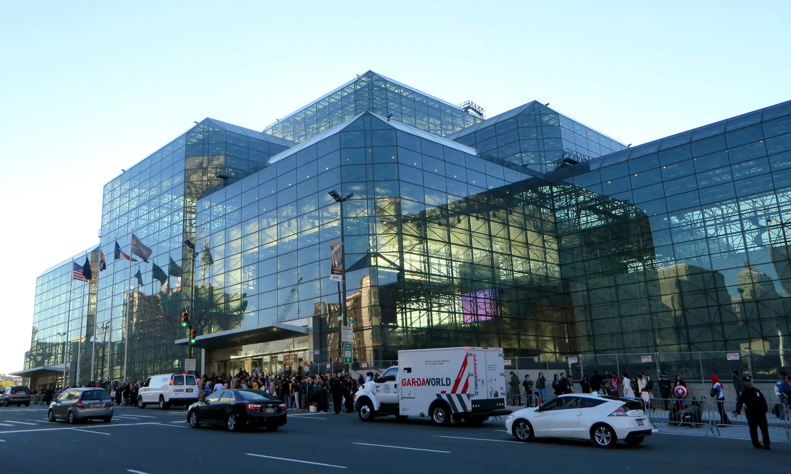 Glass exterior of the Javits Center with convention visitors and vehicles outside.