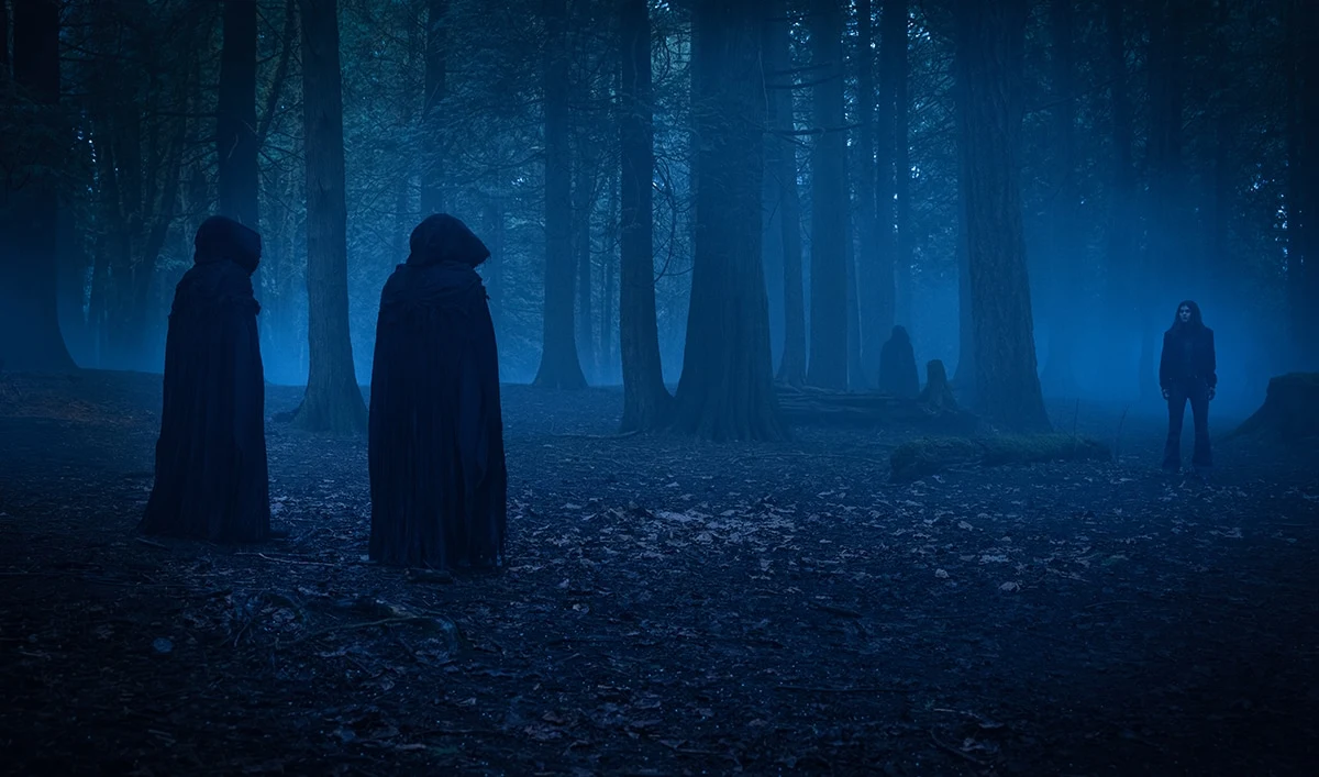 Official Mayfair Witches image showing a woman facing hooded figures in a dark blue forest.
