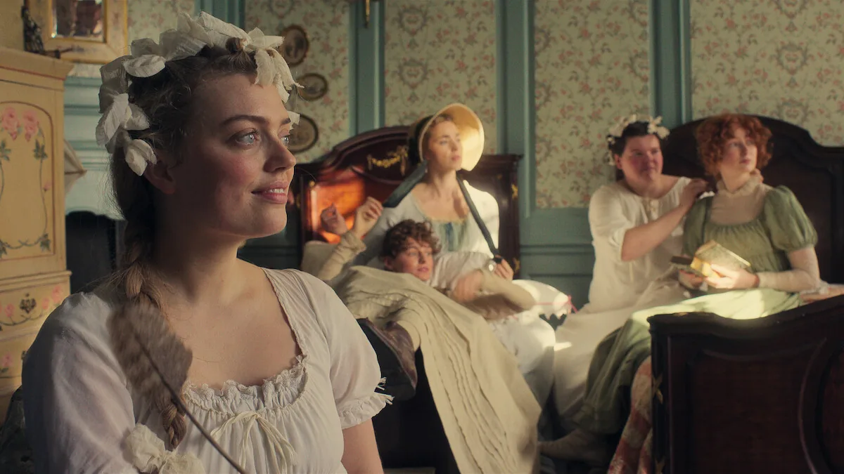 Five women in Regency dress share a floral bedroom in a still from Netflix's Pride and Prejudice, one in the foreground with a white hair ribbon.