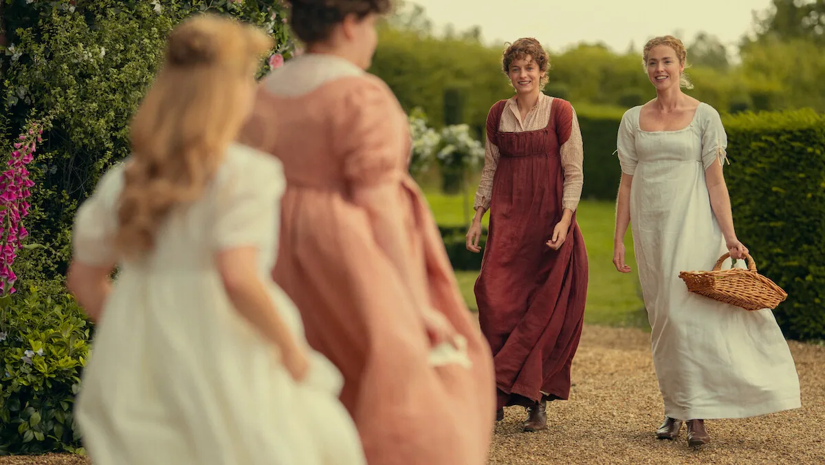 Four women in Regency dresses walk a garden path in a still from Netflix's Pride and Prejudice, two of them smiling toward the camera.