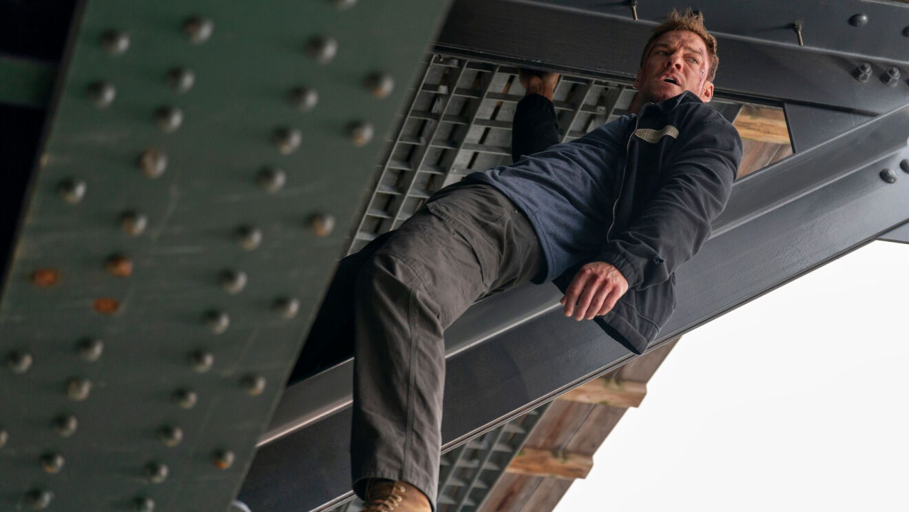 Alan Ritchson as Jack Reacher hanging beneath a steel structure in Reacher Season 4.