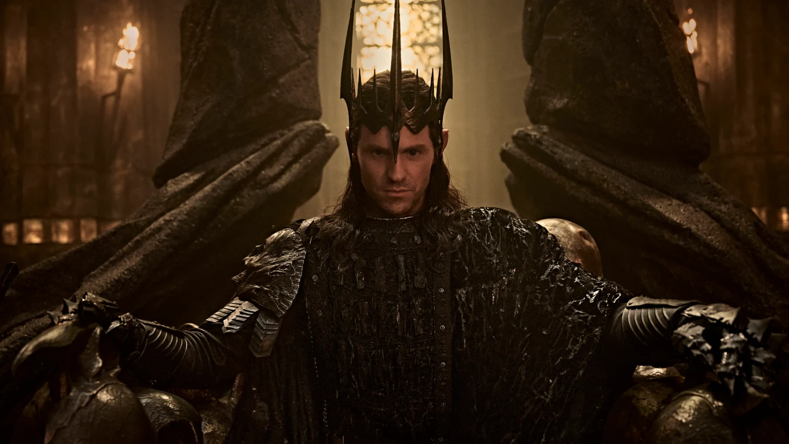 Charlie Vickers as Sauron seated in dark armor and a spiked crown in an official The Rings of Power Season 3 production still.