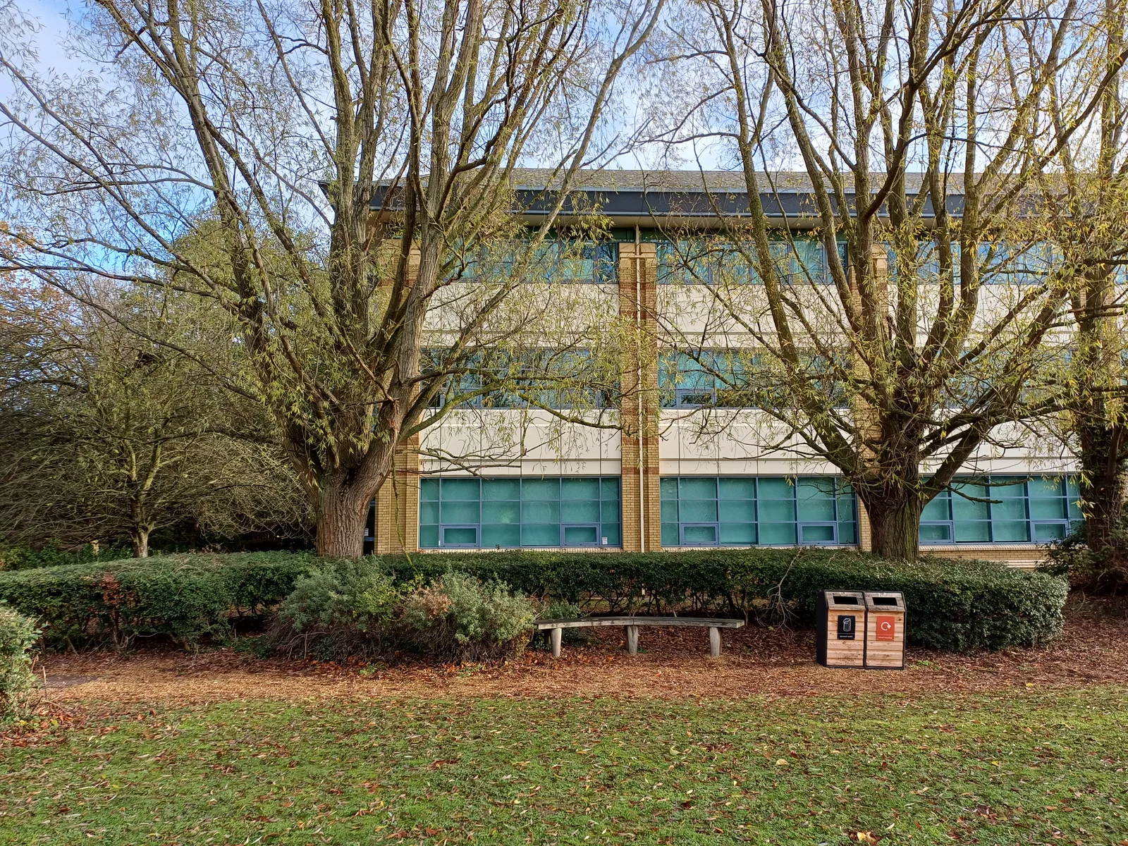 A three-storey office building at Cambridge Science Park stands behind willow trees, hedges and a bench.