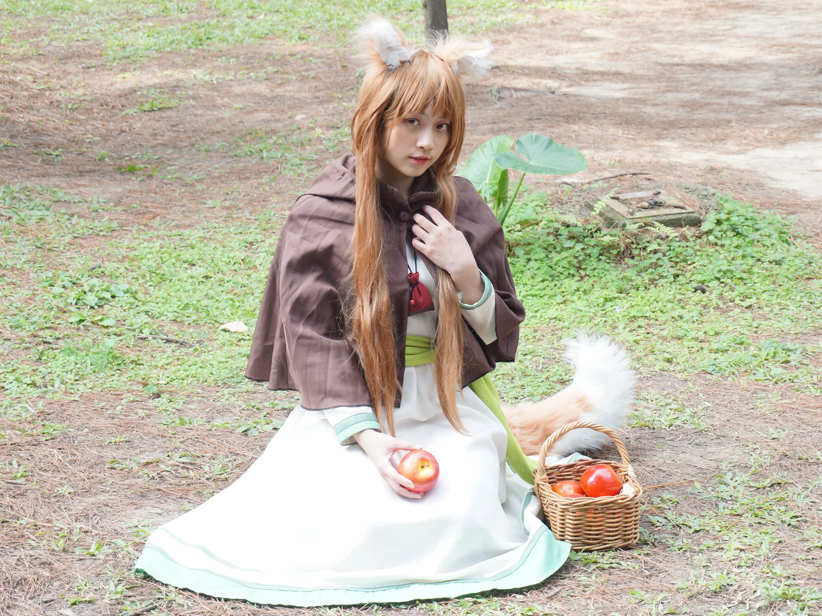 Yeyike cosplaying Holo outdoors at GJ24, wearing wolf ears and a brown cape while holding an apple beside a wicker basket.