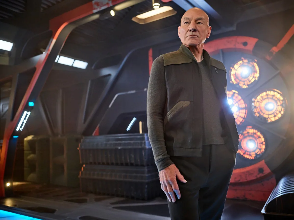 Patrick Stewart as Jean-Luc Picard standing on a dimly lit ship set in a promotional photograph for the first season of Star Trek: Picard.