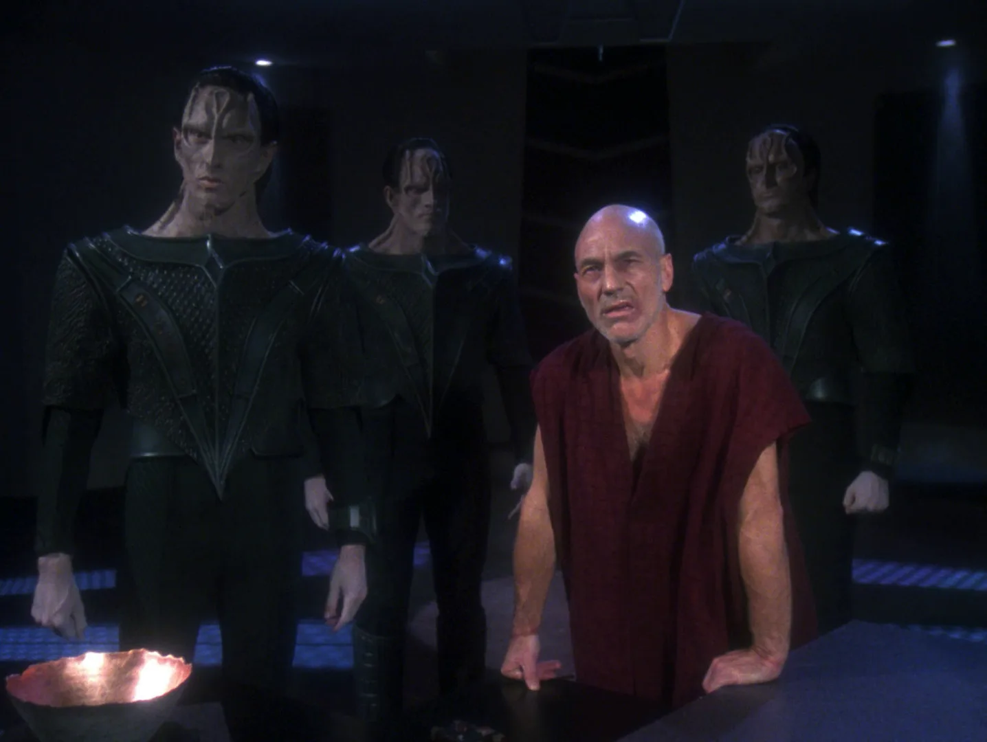 Picard, in a plain tunic, kneels defiantly among Cardassian guards in Chain of Command, Part II.