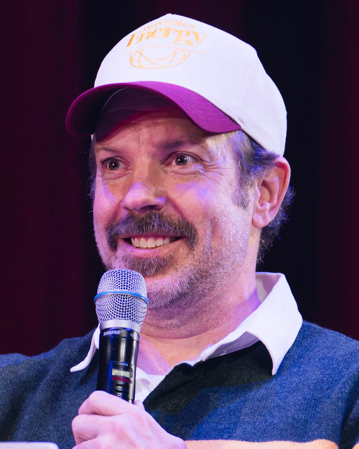 Jason Sudeikis wearing a cap and holding a microphone at a book discussion.