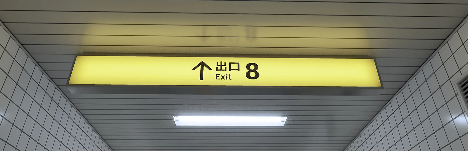 Official key art for The Exit 8, showing the game's glowing yellow subway exit sign reading 'Exit 8' inside a tiled underground passageway.