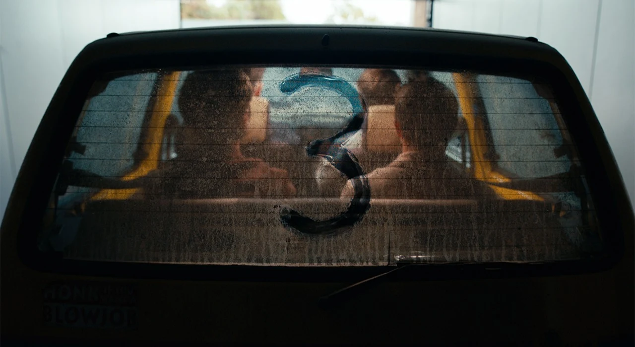 Official Netflix announcement image for The Inbetweeners 3, showing the four leads' silhouettes inside the yellow Fiat Cinquecento through its dusty rear windscreen, with a '3' drawn in the dust.