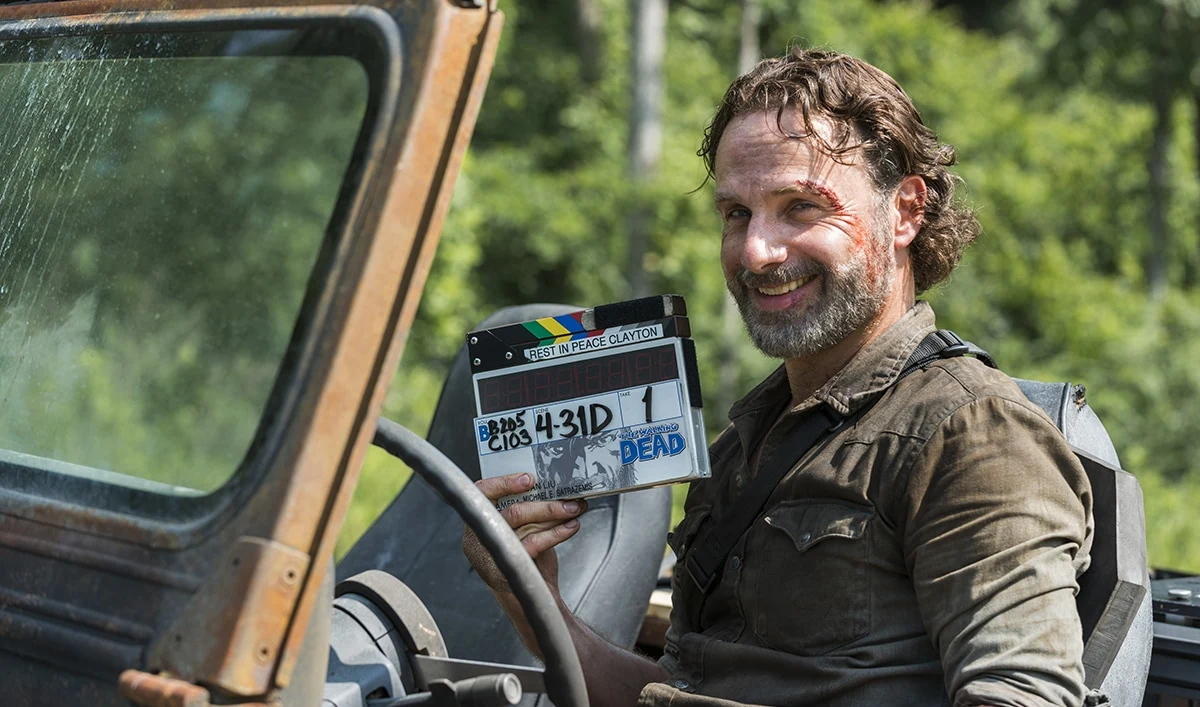 Andrew Lincoln in Rick Grimes costume smiling in a vehicle while holding a Walking Dead production clapperboard.