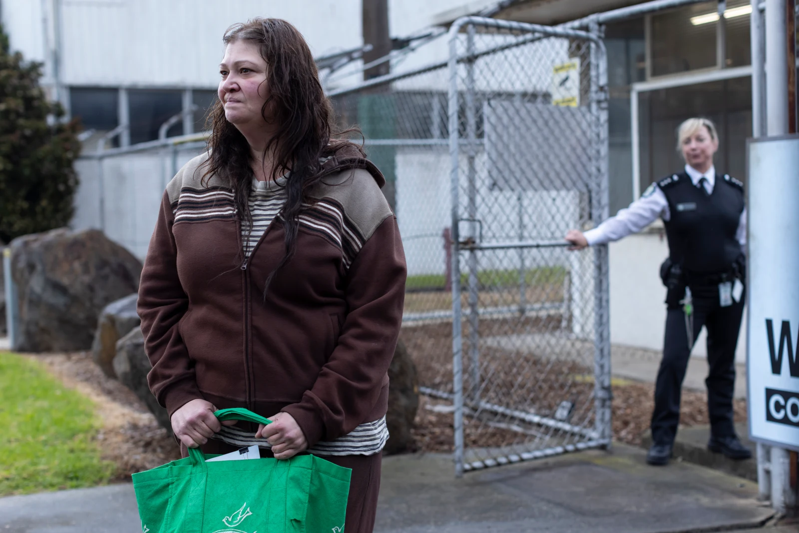 Katrina Milosevic as Boomer stands outside a prison gate holding a green bag, with Jacquie Brennan as Smiles behind her in Wentworth: Halfway Home.