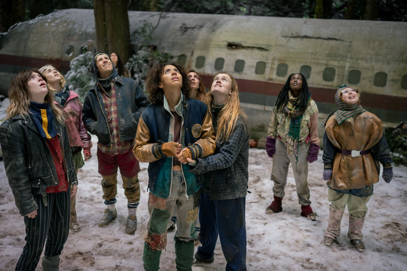 The teenage Yellowjackets ensemble looks upward beside the crashed plane in an official Season 4 production still.